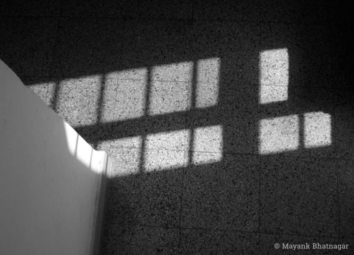 Light from a tall grilled window falling on textured floor tiles and an adjacent wall