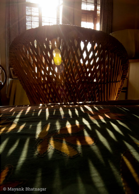 Sunlight from the window behind falling on a cane chair and its shadow patterns falling on a colourful bed cover in the foreground
