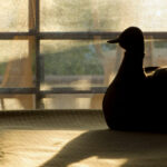 A decorative wooden duck in silhouette against light falling from the window behind on the tablecloth below