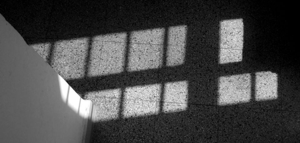 Light from a tall grilled window falling on textured floor tiles and an adjacent wall