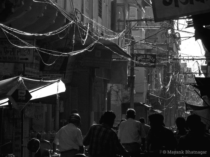 People walking below overhead wires lit-up by the sun, in an otherwise dark lane