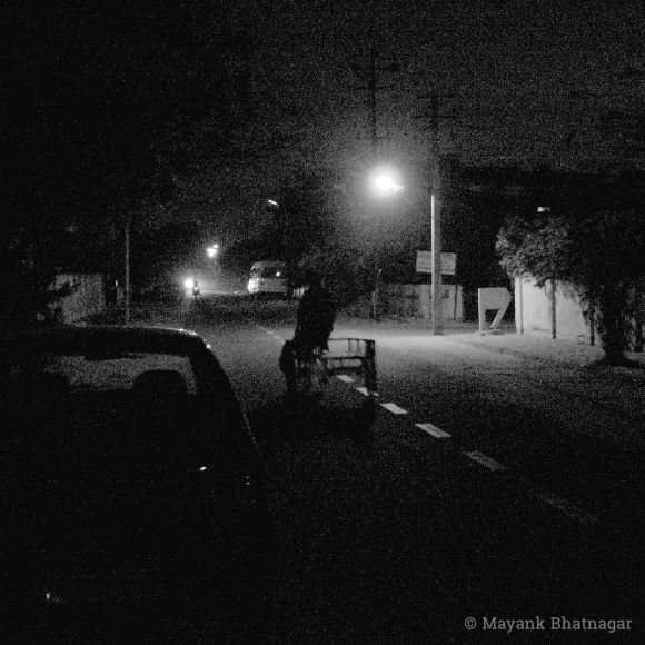 Dark, grainy photo of a cycle rickshaw in the middle of a near empty road lit up by a street light