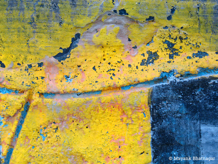 Bright yellow paint peeling off a wall with linear gashes on it, along with touches of dark blue and pink colours