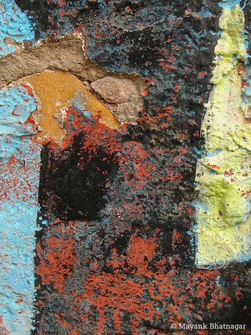 Light blue, black, red and yellow paint on a heavily textured, partly broken wall, revealing a layers of stone underneath