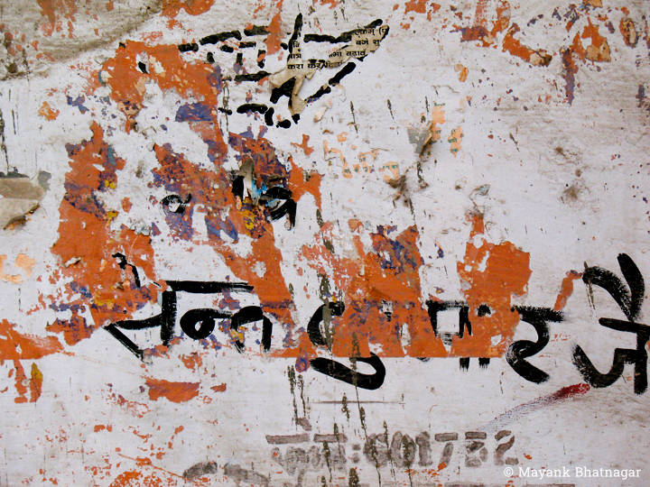 Remnants of torn red paper stuck on a white wall, over some black hand painted hindi text