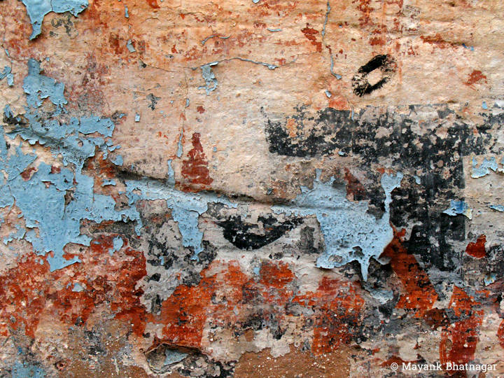 Several layers of paint: sky blue, black and red, peeling off the surface of an old, dented wall