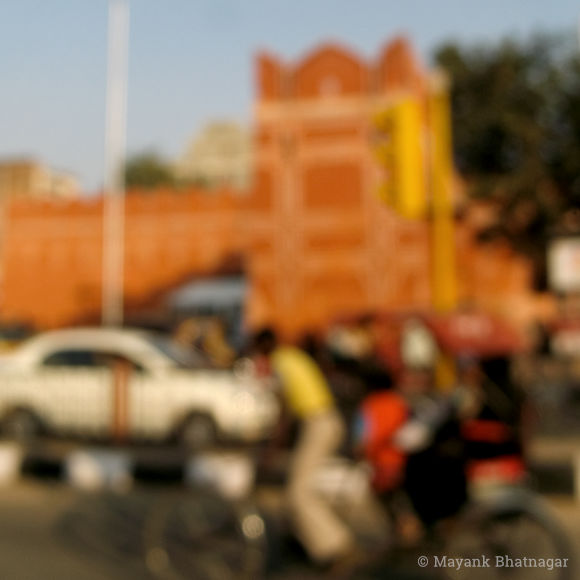 Blurred photograph of a cycle rickshaw and moving traffic in front of an old, painted city wall