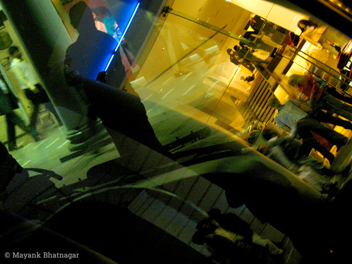 People on two levels and a shop window with mannequins, layered in a reflection on the glass of a car