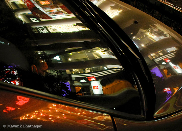 Reflections of shops and signages at night on the shiny body and windscreen of a car