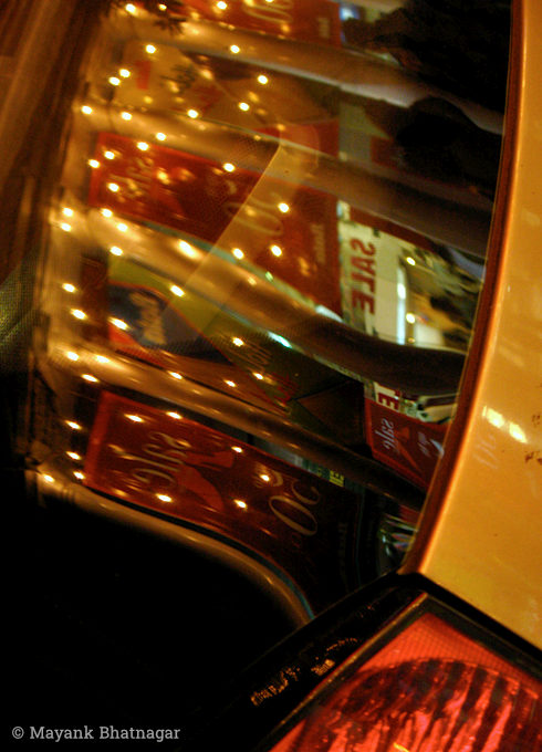 Reflections of lit up bulbs and shop signages on the rear window of a car with its tail light partly visible