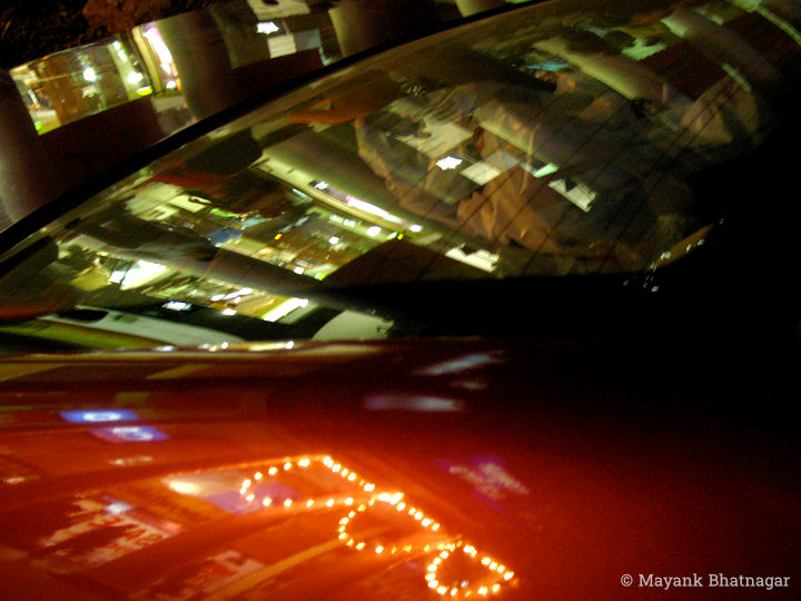 Reflections of shop lights and decorations on the windscreen and red bonnet of a car
