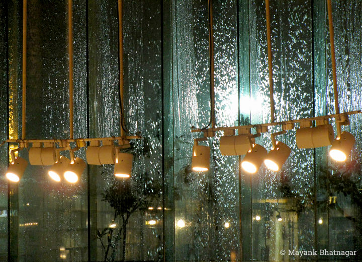 Back-lit rainwater falling on glass windows, behind lights hanging in the mall interior