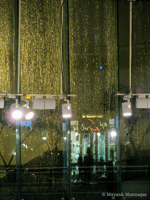 Back-lit rainwater falling on large glass windows with hanging lights and reflections of people in the foreground