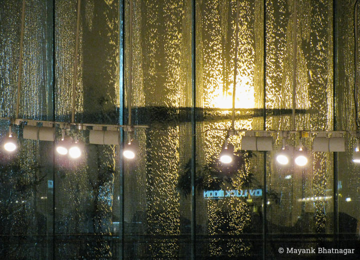 Back-lit rainwater falling on large glass windows with hanging lights and reflections of signages in the foreground