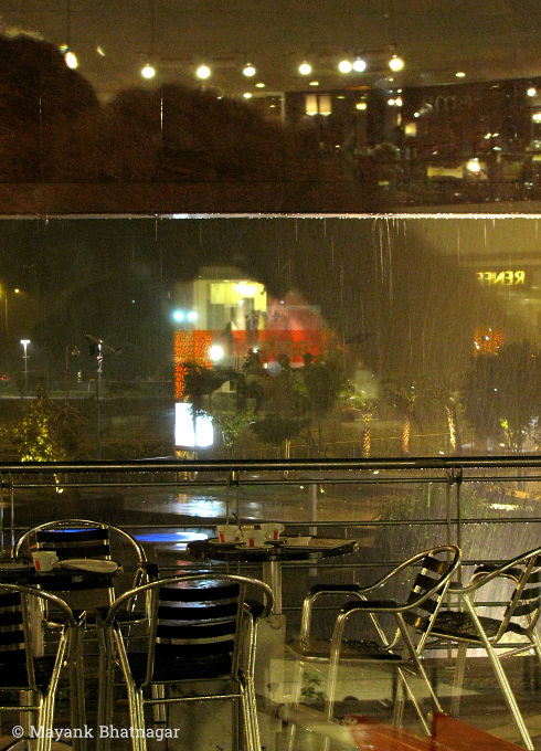 Rain falling over chairs and a railing, with buildings and street lights in the background