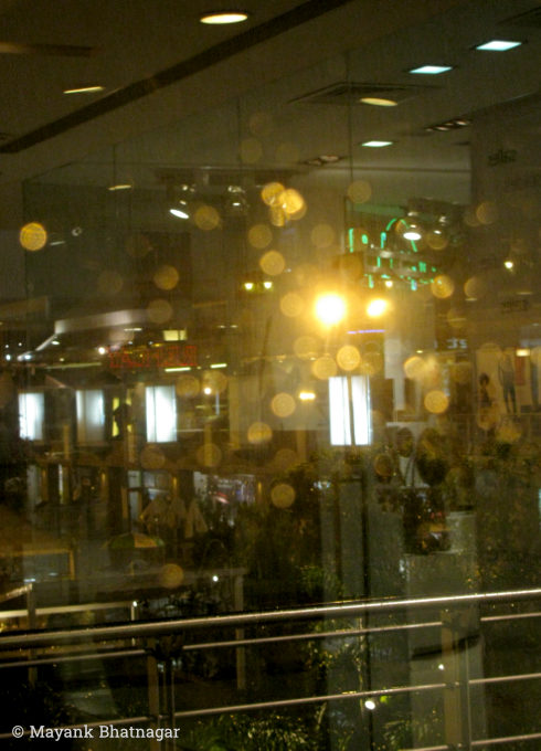 Blurred water droplets and reflections of ceiling lights on window glass, with mall exterior in the background