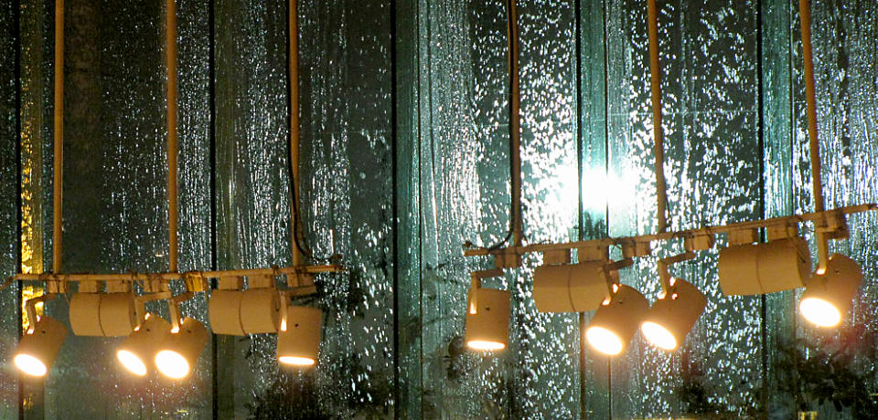 Back-lit rainwater falling on large glass windows with lights hanging in the foreground